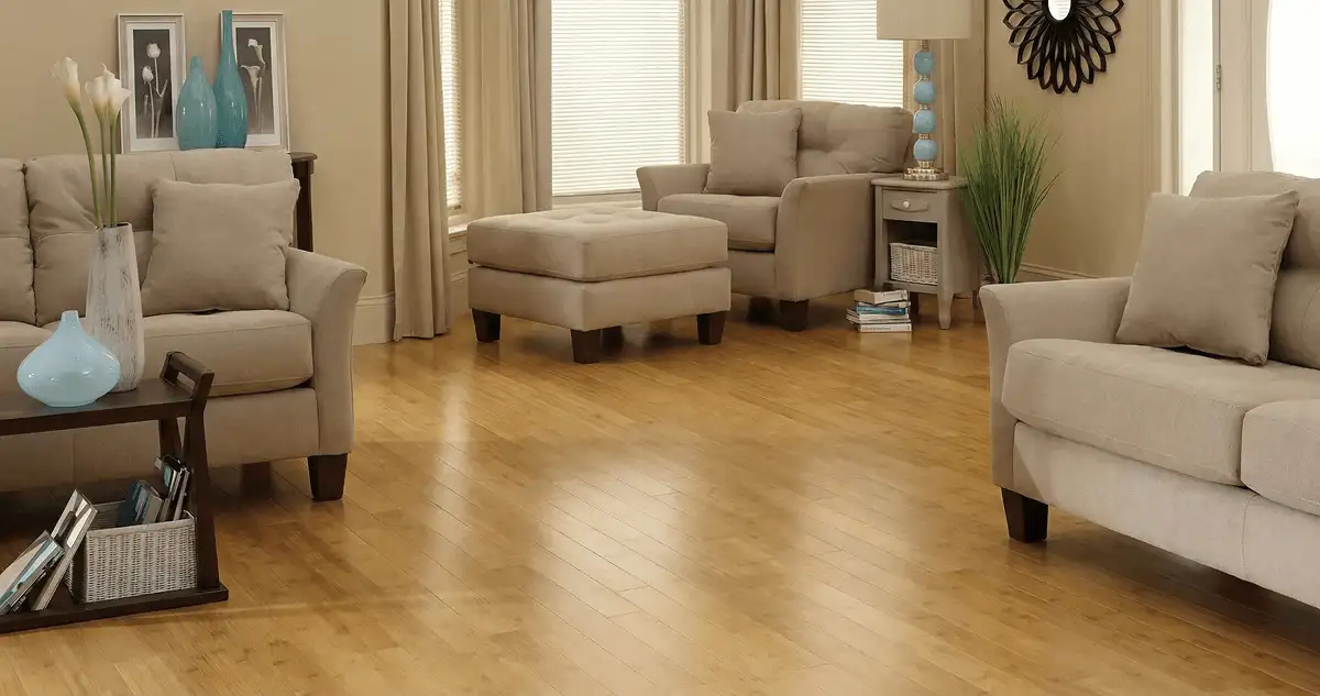 A living room with professionally installed bamboo flooring, beige sofas, a matching ottoman, a side table with a blue lamp, vases, framed pictures, and large windows with blinds and curtains in New Haven, CT